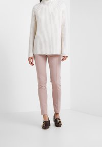 Vrouw in een witte kabelbreiselsweater, lichtroze fitted broek en donkerbruine pumps met grote gouden gespen.