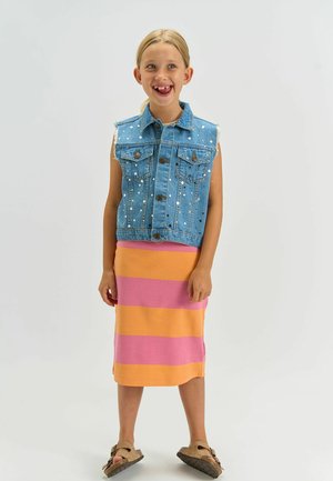 Fată tânără zâmbitoare, stând în picioare, purtând un vestă de denim albastră fără mâneci cu ținte, o fustă cu dungi roz și portocalii și sandale maro.