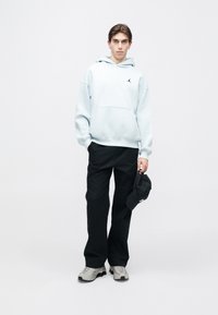 Sweatshirt bleu clair avec une poche avant et un logo noir, associé à un pantalon noir et des chaussures grises, tenant une casquette noire.