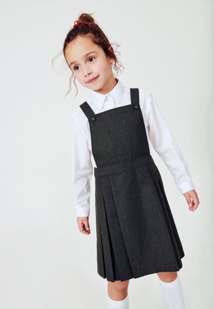 Donkergrijze tuinbroekjurk met knoopbanden over een witte blouse met kraag, voorzien van plooien en een gladde textuur, gecombineerd met witte kniekousen.