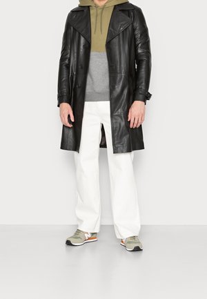 Homme portant un manteau en cuir noir par-dessus un sweat à capuche vert et gris, un pantalon blanc et des baskets vertes avec des détails orange et blanc, debout sur un sol gris.