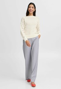 Cremefarbener gestreifter Strickpullover mit geripptem Saum, kombiniert mit hellgrauen, plissierten Hosen. Das Outfit wird durch rote Ballerinas ergänzt.