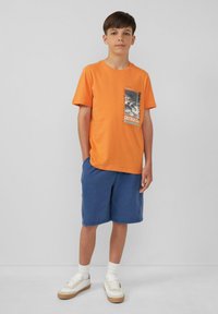 Oranje katoenen t-shirt met een grafisch zakontwerp, gecombineerd met blauwe shorts, witte sneakers en contrasterende gestreepte sokken.