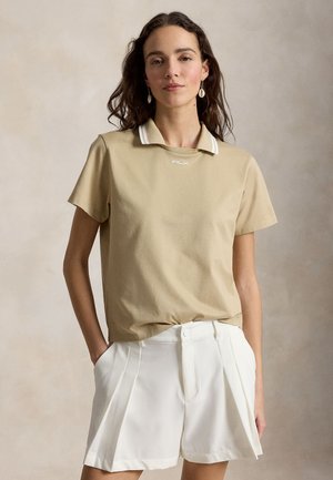 Moteris su banguotais tamsiais plaukais, dėvinti smėlio spalvos trumpomis rankovėmis polo marškinėlį ir baltus klostuotus šortus, stovinti su rankomis kišenėse prieš neutralią foną.