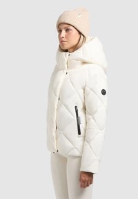 Chaqueta blanca con acolchado, cuello alto, cremalleras laterales y un detalle de botón negro. Llevada con un gorro de punto acanalado en un tono beige claro.