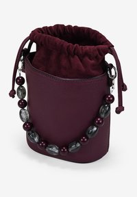 Pequeño bolso tipo cubo de cuero morado oscuro con parte superior de tela suave y cordón decorativo de cuentas en tonos morado y gris.