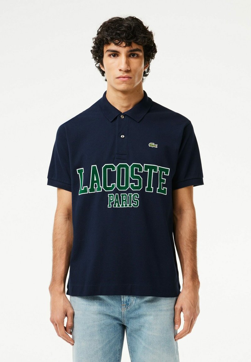 Polo-shirt bleu marine en coton avec un col classique, arborant un grand texte "LACOSTE PARIS" en blanc et en vert, ainsi qu'un petit logo vert sur la poitrine gauche.
