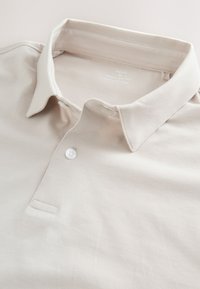Polo beige en coton avec un col à pont et un bouton blanc sur la patte de boutonnage, posé à plat pour afficher les détails.