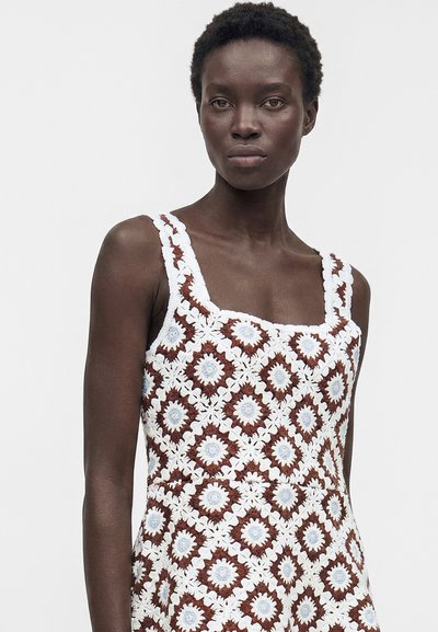 Femme aux cheveux courts et bouclés portant une robe sans manches avec des motifs carrés floraux au crochet blanc, marron et bleu clair sur un fond uni.