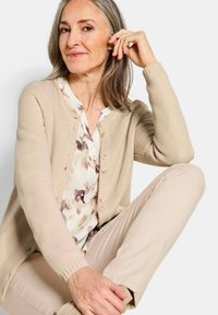 Ältere Frau mit grauem Haar, die eine beige Strickjacke, eine gemusterte Bluse und hellbeige Hosen trägt, sitzt mit einem hochgezogenen Knie vor einem weißen Hintergrund.