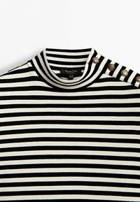 Haut en maille côtelée noir et blanc à rayures, avec col montant et quatre boutons dorés décoratifs sur l'épaule.