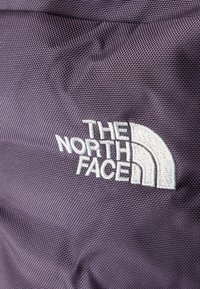 Κοντινό πλάνο μοβ υφάσματος με υφή και κεντημένο λευκό λογότυπο "THE NORTH FACE" και σχέδιο μισού θόλου.