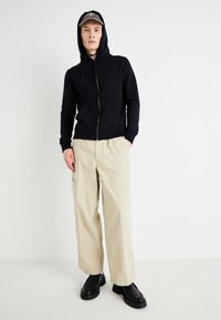 Black zip-up hoodie with a hood, beige wide-leg cargo pants with side pockets, and black chunky shoes, set against a plain white background.