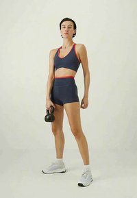 Ženska v športni opremi drži kettlebell v eni roki in samozavestno stoji na enobarvni svetli podlagi.