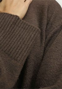 Handen vilar på axeln, iklädd en brun stickad tröja med ribbade muddar och mjukt texturerad tyg.