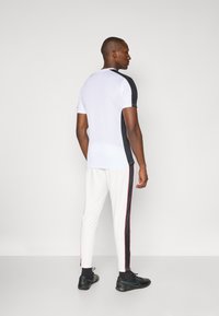 T-shirt de sport en blanc et noir, à manches courtes avec une texture en maille. Associé à un pantalon blanc avec des bandes latérales noires et roses.