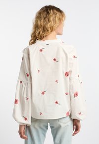 Cremefarbene Bluse mit langen, puffartigen Ärmeln, verziert mit rosa floralem Stickereien. Weicher, leichter Stoff mit einem runden Halsausschnitt und gerafften Details.