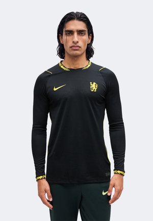 Homme aux cheveux noirs portant un maillot de sport noir à manches longues avec un logo Nike jaune et un emblème de lion, debout devant un fond uni.