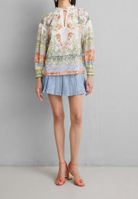 Blouse à imprimé floral avec un col noué, manches longues et poignets élastiques. Associée à une jupe plissée en denim clair et des sandales à talons orange.