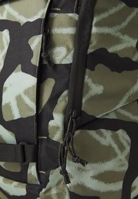 Close-up of green and black camouflage backpack fabric with two black zipper pulls and a black strap.