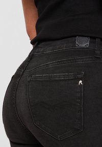 Vaqueros de mezclilla negra con un diseño ajustado, que presentan un bolsillo trasero con un acento plateado, detalles de costura sutiles y un tejido texturizado.