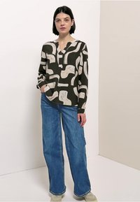 Camisa de manga larga en verde oscuro con patrones abstractos en color crema; combinada con vaqueros azul sueltos y zapatillas grises.