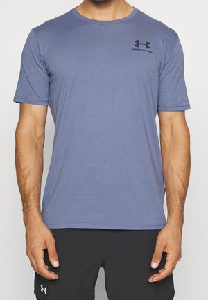 Mężczyzna w niebieskiej koszulce Under Armour z krótkim rękawem i czarnych szortach, stojący z rozluźnionymi ramionami wzdłuż ciała.