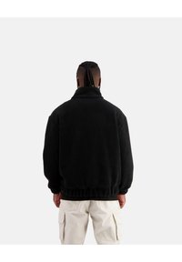 Schwarze Fleecejacke mit hohem Kragen, strukturiertem Material, elastischem Saum und lockerer Passform. Kombiniert mit hellen Cargo-Hosen.