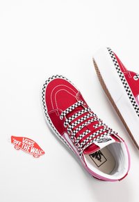 Baskets montantes rouges Vans avec des lacets et des bordures à damier noir et blanc, présentées avec une étiquette logo Vans "Off The Wall" sur fond blanc.