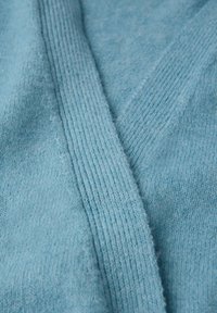 Ljusblå stickad kofta med V-ringning, ribbad struktur och mjukt utseende. Närbild som fokuserar på tygdetalj och veck.