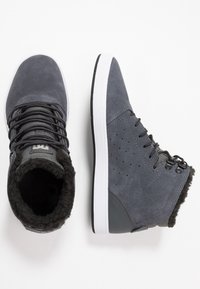 Harmaat mokkasiinimaiset korkeat lenkkitossut, joissa on mustat nauhat, fleecevuori, valkoinen kumipohja ja rei'itetyt yksityiskohdat sivuilla.