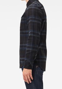 Camisa de cuadros en negro y azul, de manga larga y con botones, presenta un cuello, un bolsillo en el pecho y un dobladillo recto. La tela parece suave y texturizada.