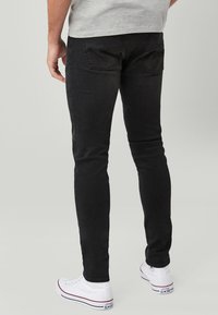 Crne skinny traperice izrađene od denima, s glatkom teksturom i stražnjim džepovima. Uparene s bijelim tenisicama, ističu ležeran izgled.