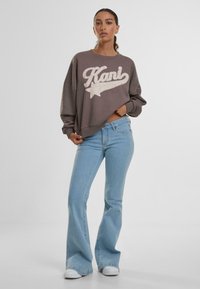Sudadera corta marrone con testo "Kani" crema e grafica di stella. Abbinata a jeans chiari a campana e sneaker bianche.