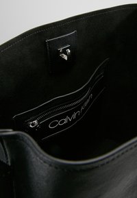 Wnętrze czarnej zamszowej torby typu tote, z przegrodą zapinaną na zamek i metką "Calvin Klein" w białym kolorze, srebrne detale okucia na klapie.