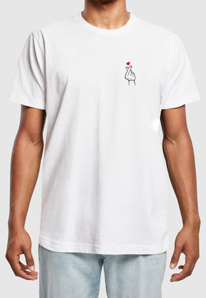 Biała koszulka noszona przez osobę, z małą dłonią robiącą gest serca z palców, z czerwonym sercem nad nią, na klatce piersiowej.