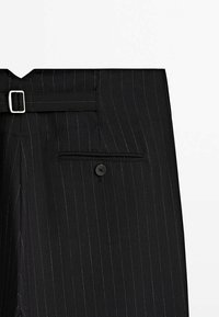 Pantalon noir à fines rayures avec une boucle latérale et une poche arrière passepoilée avec un seul bouton sur fond blanc.
