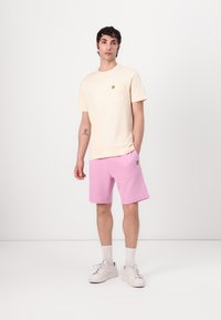 Ljusbeige t-shirt med kort ärm och bröstficka, liten gul logga, matchas med rosa shorts och vita sneakers. Enkelt design, bomullsmaterial.