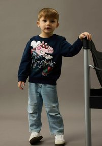 Niño pequeño con un suéter azul marino que tiene un diseño de un cerdo de dibujos animados y una ciudad, jeans azul claro, zapatillas blancas, de pie y sosteniendo una silla.