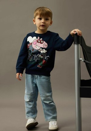 Niño pequeño con un suéter azul marino que tiene un diseño de un cerdo de dibujos animados y una ciudad, jeans azul claro, zapatillas blancas, de pie y sosteniendo una silla.