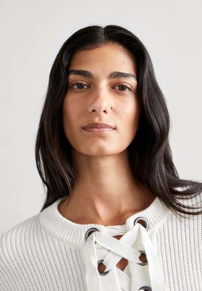 Witte gebreide top met een ronde halslijn en vetersluiting met zilveren eyelets. De textuur is geribbeld, wat visuele interesse toevoegt.