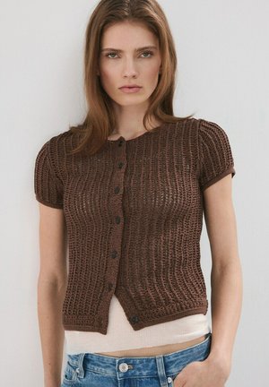 Mujer joven con cabello castaño liso, vestida con un cárdigan marrón de punto de manga corta abotonado sobre una camiseta blanca y jeans azules.