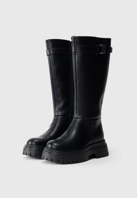Botas de couro pretas até ao joelho com sola de tração grossa, fecho lateral e uma fivela decorativa na tira superior. Textura suave.