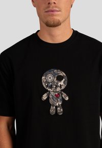 Černé tričko s vyšitým nášivkem kreslené postavičky s texturovaným designem, který zahrnuje modré, stříbrné a červené akcenty.