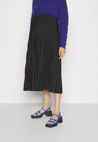 Gonna nera plissettata abbinata a un maglione blu e sandali blu con cinturini, dotati di un tacco nero spesso. Tessuto liscio e design fluido.