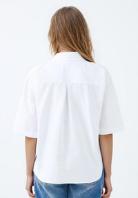 Weißes Kurzarmhemd mit Kragen, geradem Schnitt, Rückenschlitzdetail und glatter Textur. Kombiniert mit hellblauen Jeans.