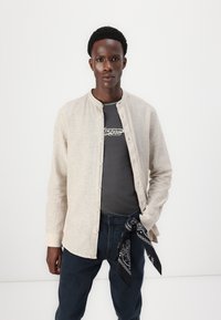 Camisa de botón beige claro con cuello de banda, combinada con una camiseta gris oscuro y jeans azul marino. Pañuelo negro atado a la cintura.