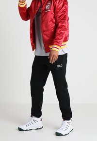 Červená saténová bomber bunda s žebrovanými manžetami, pod ní šedé tričko, černé tepláky a bílé tenisky se zelenými akcenty. Viditelné detaily loga.