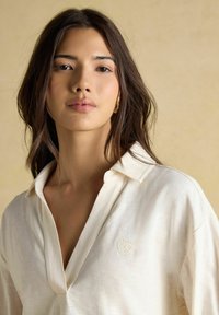 Noor juustega noor naine valges krae särgis, seistes beeži taustaga, vaatab otse ette neutraalse ilmega.