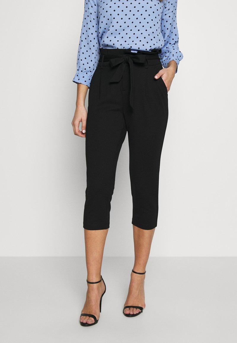 Pantalon noir court avec ceinture à nouer, en tissu lisse ; porté avec un chemisier bleu clair à pois et des talons noirs à brides.